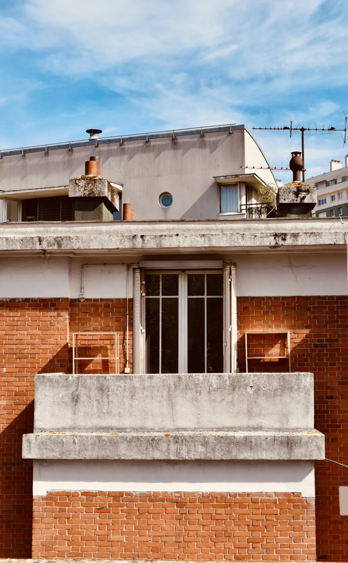 Deniz · 06/09/2025 · 3551 x 5735 Cette photographie met en scène un bâtiment aux allures industrielles avec une façade en brique rouge et des éléments en béton brut. Le sujet principal est un balcon en saillie, partiellement caché par une structure en béton.La composition joue sur les lignes horizontales et verticales, créant un contraste géométrique. Le cadrage est serré, centré sur les détails architecturaux.La lumière naturelle, vive et contrastée, accentue les textures et les ombres. Les couleurs dominantes sont les tons chauds des briques et les gris du béton, avec des touches de bleu dans le ciel.L’ambiance est à la fois austère et minimaliste, évoquant une architecture moderne et brute.