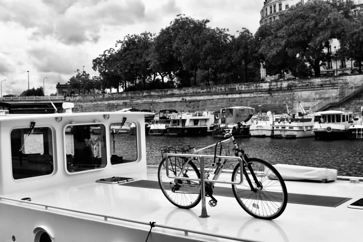 Deniz · 13/09/2025 · 6000 x 4000 Cette photographie en noir et blanc met en scène un vélo attaché à la proue d'une péniche, sujet principal. La composition joue sur la juxtaposition entre l'élément urbain (le vélo) et le cadre fluvial et architectural. Le cadrage est serré sur le vélo, mais inclut aussi l'arrière-plan avec des bateaux et des bâtiments.La lumière semble naturelle, avec des contrastes marqués. Les couleurs dominantes, bien que monochromes, varient du noir au blanc en passant par des gris. Le style est documentaire et réaliste, évoquant une ambiance paisible et légèrement nostalgique.