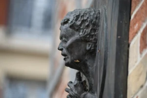 Pierre · 22/11/2025 · 6048 x 4024 Cette photographie met en scène une **sculpture en bronze** représentant une tête féminine stylisée, aux traits flous et expressifs. Le **cadrage serré** isole le sujet, accentuant ses détails et textures.La **composition** joue sur le contraste entre la sculpture et le fond flou, avec une **lumière naturelle** douce qui crée des ombres légères. Les **couleurs dominantes** sont les tons **noirs et gris** de la sculpture, contrastant avec les teintes chaudes et floues du bâtiment en arrière-plan.Le style évoque une **artistique abstraction**, tandis que l’ambiance, à la fois mystérieuse et contemplative, suggère une touche poétique et intemporelle.