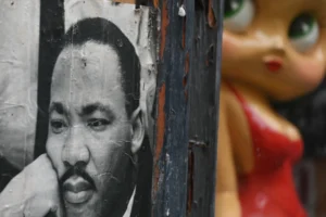 Pierre · 22/11/2025 · 3808 x 2533 Cette photographie présente deux sujets principaux : un portrait délavé de Martin Luther King Jr. et une poupée aux traits stylisés, aux couleurs vives (rouge et bleu).La composition juxtapose ces éléments contrastés. Le cadrage serré isole les visages, créant une tension visuelle. La lumière semble naturelle mais tamisée, accentuant les dégradés et les textures.Les couleurs dominantes sont les tons sépia du portrait et les couleurs vives de la poupée. Le style mêle réalisme et naïf, évoquant une ambiance à la fois mélancolique et nostalgique, voire critique.
