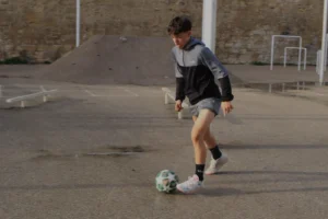 Marie-Odile · 1600 x 1066 Cette photographie capture un jeune garçon en action, sujet principal, jouant au football. La composition montre une scène dynamique avec le ballon au premier plan et le joueur en mouvement.Le cadrage est serré, mettant en valeur ses pieds et son engagement physique. La lumière semble naturelle, probablement en extérieur, avec des ombres douces. Les couleurs dominantes sont neutres (gris du sol, vert du ballon) et quelques touches vives (tenue du joueur).Le style est simple et spontané, évoquant un moment authentique. L’ambiance est sportive et joyeuse, reflétant l’énergie et la passion du jeu.