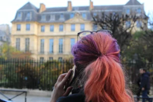 Marie-Odile · 1600 x 1066 Cette photographie met en scène une personne avec une chevelure aux reflets **roses et violets**, attachée en un chignon haut. Le **sujet principal** est flou, attirant l’attention sur ses cheveux colorés.La **composition** est simple, avec un **cadrage serré** sur la tête, tandis qu’en arrière-plan, un **bâtiment classique** et des arbres se distinguent. La **lumière** semble naturelle, douce et légèrement tamisée, créant une ambiance calme.Les **couleurs dominantes** sont les tons pastel des cheveux et les teintes neutres du bâtiment. Le style est **minimaliste et spontané**, évoquant une **ambiance urbaine et artistique**.