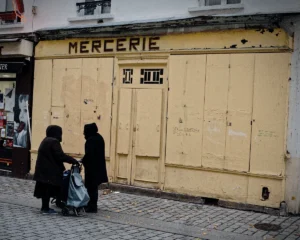 Cyrille · 22/11/2025 · 1200 x 960 Cette photographie montre deux femmes voilées devant une boutique fermée nommée "Mercerie". Le sujet principal est leur silhouette, contrastant avec la porte jaune fermées et barricadée.La composition est simple et symétrique, avec un cadrage serré sur les femmes et la boutique. La lumière est naturelle, tamisée, créant des ombres douces. Les couleurs dominantes sont les tons jaunes et beiges de la boutique, avec des noirs et gris des vêtements des femmes.Le style est documentaire et réaliste, évoquant une ambiance calme et mélancolique, presque urbaine et intemporelle.