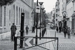 Cyrille · 22/11/2025 · 1200 x 798 Cette photographie en noir et blanc capture une rue urbaine étroite et légèrement pentue. Le sujet principal semble être l’architecture et l’ambiance de la rue, avec des bâtiments anciens en arrière-plan. La composition met en valeur des lampadaires alignés et un panneau en forme de "K" au premier plan.Le cadrage est serré, centré sur la perspective des passants et des détails architecturaux. La lumière est naturelle, avec des contrastes marqués créant des ombres nettes. Les couleurs dominantes, bien que monochromes, varient du noir au gris clair.Le style est documentaire et réaliste, évoquant une ambiance calme et légèrement mélancolique.