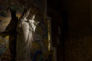 Cyrille · 22/11/2025 · 1200 x 798 Cette photographie met en scène une statue religieuse, probablement une Vierge à l'Enfant, éclairée par une lumière douce et directionnelle. Le sujet principal est centré, avec un cadrage serré qui isole la figure.La lumière, contrastée, accentue les détails et crée une ambiance mystique. Les couleurs dominantes sont les teintes chaudes et dorées de l'éclairage, contrastant avec les tons sombres et les nuances bleutées de l'arrière-plan.Le style évoque l'art sacré, avec des éléments décoratifs peints sur les murs. L'ambiance est solennelle et contemplative, renforcée par l'obscurité environnante.