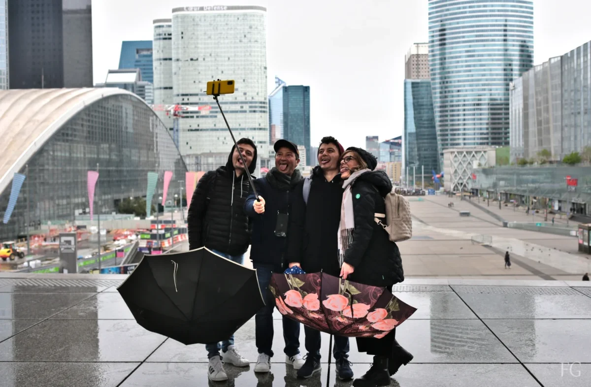 Frédéric · 23/04/2023 · 1649 x 1080 Cette photographie montre quatre personnes posant devant un bâtiment moderne aux formes arrondies, probablement un musée ou une salle de concert. Le sujet principal est ce groupe, souriant et détendu, avec un selfie stick et un parapluie.La composition est symétrique, avec un cadrage serré sur les sujets et un arrière-plan urbain. La lumière est naturelle, légèrement voilée, créant une ambiance douce. Les couleurs dominantes sont les tons froids (gris, bleu) avec des touches de noir et blanc. Le style est documentaire et spontané, évoquant une ambiance joyeuse et moderne dans un cadre urbain dynamique.