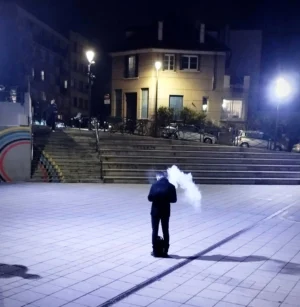 Cette photographie capture un homme en train de marcher ou de courir dans une zone enneigée, vêtu d’un manteau sombre. Le sujet principal est flou, suggérant un mouvement rapide.La composition montre un escalier de ville en arrière-plan, éclairé par des lampadaires. Le cadrage est serré sur l’individu, avec une ambiance nocturne.Les couleurs dominantes sont les tons froids : blanc de la neige, gris et bleu nuit. La lumière artificielle crée des contrastes nets. Le style est réaliste et spontané, évoquant une ambiance mystérieuse et solitaire.
