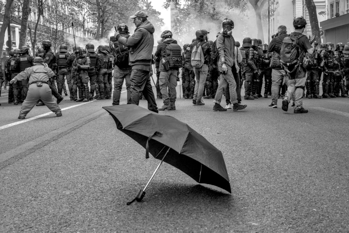 Frédéric · 01/05/2024 · 2519 x 1680 Cette photographie en noir et blanc montre une scène de manifestation. Le sujet principal est un parapluie renversé au premier plan, symbolisant peut-être la chute ou la résistance. La composition oppose ce parapluie à un groupe de policiers en formation, créant un contraste entre fragilité et autorité. Le cadrage serré et bas met en valeur cet élément central.La lumière est diffuse, renforçant l’ambiance dramatique. Les tons de gris dominent, accentuant les contrastes et les textures. Le style est documentaire et réaliste, évoquant une tension palpable et une ambiance de conflit social.