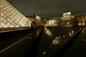 Cette photographie met en scène le **Pyramide du Louvre**, vue depuis son intérieur vitré. Le **sujet principal** est la structure géométrique lumineuse, reflétée dans le sol noir et poli.La **composition** joue sur les lignes droites et la symétrie, renforcée par le cadrage serré. La **lumière**, douce et dorée, éclaire la pyramide, créant un contraste avec l’obscurité environnante. Les **couleurs dominantes** sont les tons chauds (or) et les noirs profonds, renforçant une **ambiance mystérieuse et élégante**.Le style est **minimaliste et architectural**, évoquant modernité et grandeur historique.
