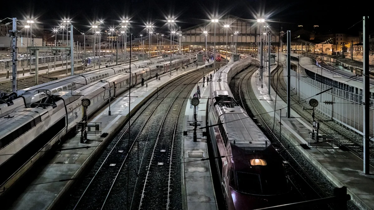 Frédéric · 06/11/2023 · 1920 x 1080 Cette photographie montre un **gare ferroviaire nocturne**. Le **sujet principal** est un train en mouvement sur des voies multiples.La **composition** est symétrique, avec des rails parallèles convergeant vers l’horizon. Le **cadrage** est large, offrant une vue aérienne. La **lumière**, artificielle et froide, provient des éclairages publics et des phares du train, créant des contrastes nets. Les **couleurs dominantes** sont des tons sombres (noirs, gris) avec des éclats jaunes et blancs.Le **style** est réaliste et documentaire, évoquant une **ambiance industrielle et dynamique**, presque solennelle.