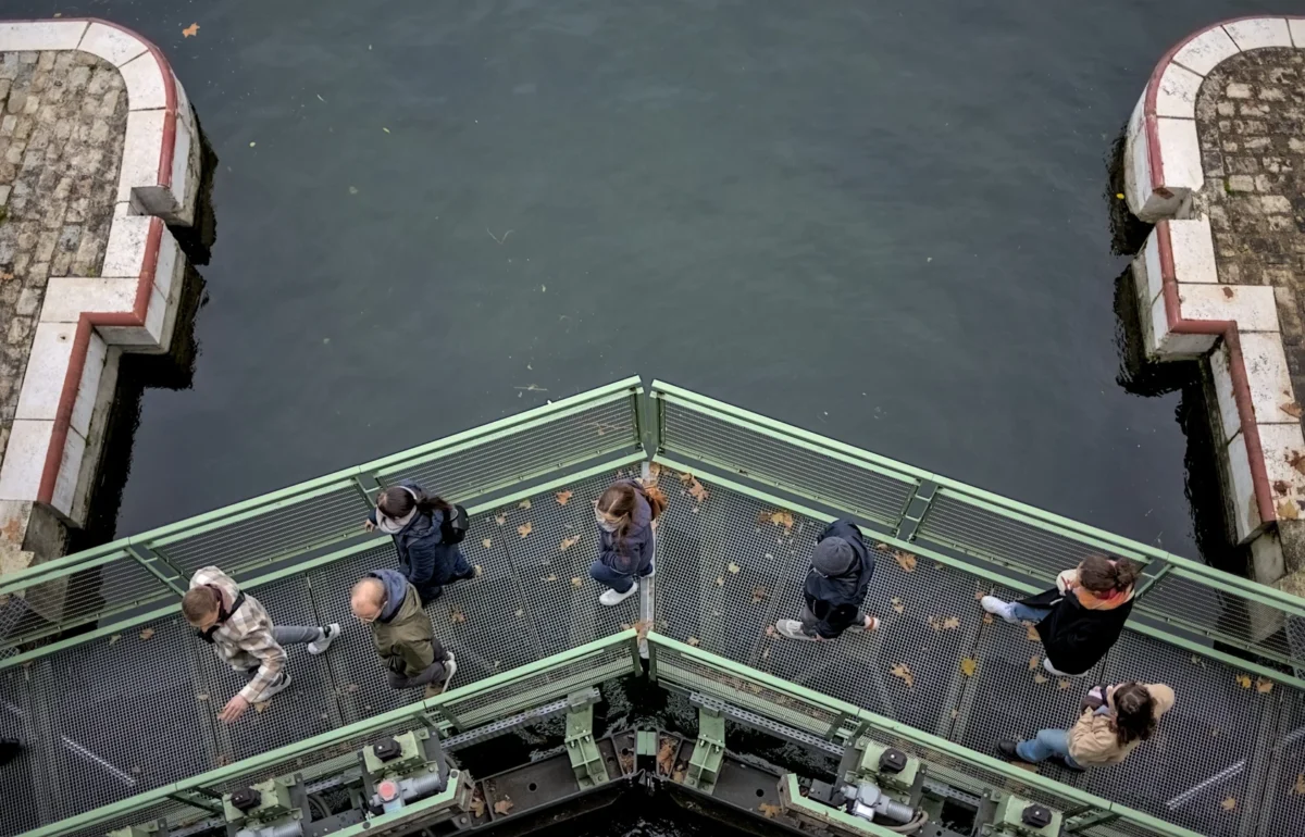 Frédéric · 11/11/2023 · 1683 x 1080 Cette photographie montre un groupe de personnes sur un pont ou une passerelle métallique enjambant un plan d’eau. Le **sujet principal** est l’assemblée de silhouettes, dont certaines semblent discuter ou observer.La **composition** est symétrique, avec un cadrage en plongée accentuant la perspective. Les **couleurs dominantes** sont les tons grisâtres du métal et de l’eau, contrastés par des éléments blancs (barrières) et des vêtements sombres.La **lumière** semble naturelle mais diffuse, créant une ambiance sobre et légèrement mélancolique. Le **style** est documentaire, avec une touche d’abstraction due à la perspective. L’**ambiance** est calme, presque introspective.
