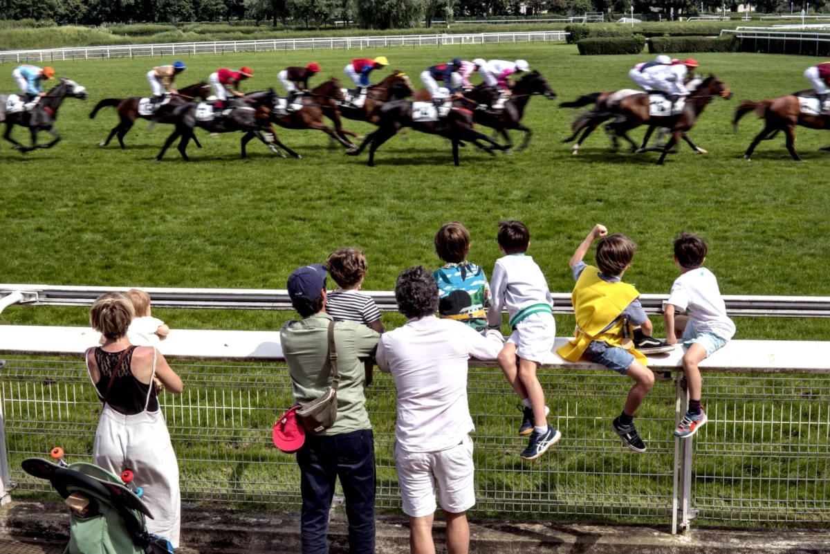 Frédéric · 08/06/2024 · 2517 x 1680 Cette photographie capture une course hippique. Le **sujet principal** est la compétition équestre en arrière-plan, avec des cavaliers et chevaux en action.La **composition** montre un groupe de spectateurs en premier plan, séparés de la piste par une barrière. Le **cadrage** est dynamique, mettant en valeur l'action des chevaux et l'attention des spectateurs.La **lumière** semble naturelle, avec des ombres douces, suggérant un jour ensoleillé. Les **couleurs dominantes** sont les verts de la piste et des arbres, les tons blancs et noirs des chevaux, et les couleurs vives des vêtements des spectateurs.Le **style** est documentaire et spontané, capturant un moment authentique. L’**ambiance** est excitante et conviviale, mêlant passion pour les courses et loisirs.