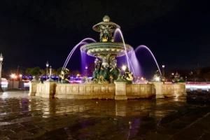 Cette photographie met en scène une **fontaine monumentale** comme sujet principal, illuminée la nuit. La composition est symétrique, avec des jets d’eau s’élevant en arcs lumineux.Le cadrage est serré sur la fontaine, intégrant partiellement l’environnement urbain en arrière-plan. La lumière, douce et artificielle, met en valeur les détails sculpturaux et crée des reflets sur le sol mouillé.Les **couleurs dominantes** sont les bleus des éclairages et les tons froids de la nuit, contrastant avec les teintes chaudes des pierres. Le style est **classique et élégant**, évoquant une ambiance **romantique et paisible**, malgré l’animation urbaine suggérée.