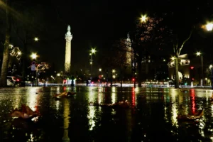 Cette photographie capture une place urbaine nocturne. Le **sujet principal** est un **monument éclairé**, probablement une colonne, reflété dans une flaque d’eau au premier plan. La **composition** est symétrique, avec des jeux de lumière et des reflets sur le sol mouillé.Le **cadrage** est large, incluant des bâtiments et des lumières festives. Les **couleurs dominantes** sont les teintes froides (noir, gris) contrastées par des éclats chauds (jaunes, rouges). L’**ambiance** est mystérieuse et poétique, renforcée par les **lumières artificielles** et les reflets aquatiques. Le style évoque une **nuit animée et magique**.