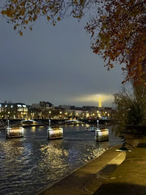 Cette photographie capture un **canal urbain nocturne**. Le **sujet principal** est le reflet des lumières sur l’eau et les bateaux amarrés.La **composition** joue sur les lignes horizontales du quai et de l’eau, encadrée par des arbres aux feuilles automnales. Le **cadrage** est serré sur le premier plan, avec une perspective légèrement inclinée.Les **lumières** douces et artificielles créent une ambiance tamisée, tandis que les **couleurs dominantes** sont des teintes bleutées et dorées, accentuées par les éclairages urbains.Le **style** est réaliste et contemplatif, évoquant une **ambiance calme et poétique**, typique d’une ville européenne en soirée.