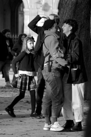 Thomas · 01/11/2025 · 1080 x 1624 Cette photographie en noir et blanc met en scène un groupe de personnes en extérieur. Le sujet principal semble être un homme au centre, tenant un appareil photo et observant à travers le viseur, entouré de deux femmes.La composition est dynamique, avec des lignes de fuite vers l’arrière-plan. Le cadrage est serré sur les personnages, mais inclut aussi des éléments du décor urbain. La lumière est douce, créant des contrastes marqués et des ombres nettes. L’absence de couleurs accentue les textures et les jeux de lumière.L’ambiance est à la fois quotidienne et intemporelle, évoquant une scène de rue authentique et spontanée.