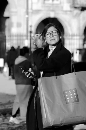 Thomas · 01/11/2025 · 1080 x 1625 Cette photographie en noir et blanc met en scène une femme comme sujet principal, tenant un grand sac à provisions. Elle semble observer quelque chose avec attention, peut-être son téléphone.La composition est simple et centrée, avec un cadrage serré sur la femme, légèrement floue en arrière-plan. La lumière est douce, naturelle, créant des contrastes modérés. L’ambiance est urbaine et quotidienne, renforcée par les silhouettes floues en arrière-plan.Le style est documentaire, réaliste, capturant un moment authentique. Les tons de gris dominent, accentuant les détails et les textures.