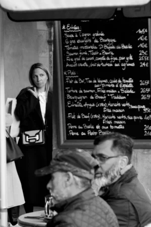 Thomas · 01/11/2025 · 1080 x 1625 Cette photographie en noir et blanc capture une scène intimiste dans un restaurant. Le sujet principal est une femme debout près d’un tableau de menu écrit à la craie, observant deux hommes assis à une table.La composition joue sur les contrastes entre l’espace intérieur et les regards. Le cadrage serré met en valeur les expressions et les détails des visages. La lumière douce, probablement naturelle, accentue les ombres et crée une ambiance chaleureuse et nostalgique. Les tons gris dominent, renforçant l’aspect rétro et authentique de la scène, évoquant une ambiance simple et conviviale des années passées.