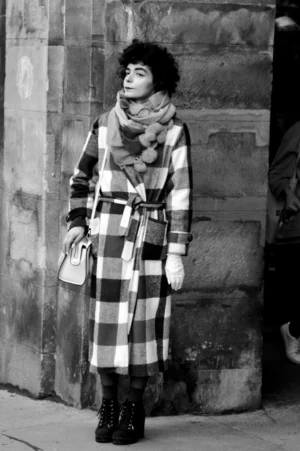Thomas · 01/11/2025 · 1080 x 1625 Cette photographie en noir et blanc met en scène une personne comme sujet principal, vêtue d’un long manteau à carreaux et d’un écharpe, posant devant un mur texturé.Le cadrage est serré, centré sur elle, avec une composition verticale. La lumière semble naturelle, douce et légèrement contrastée, accentuant les détails du vêtement et du visage.Les couleurs, bien que monochromes, jouent sur les variations de gris et de noir, créant une ambiance rétro et élégante. Le style évoque un classicisme intemporel, renforcé par le jeu des textures et des motifs. L’ambiance est à la fois sobre et chaleureuse.