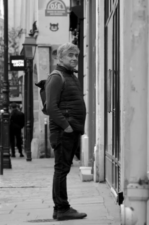 Thomas · 01/11/2025 · 1080 x 1625 Cette photographie en noir et blanc met en scène un homme comme sujet principal, debout près d’un mur, les mains sur les hanches, vêtu d’un blouson et d’un pantalon. La composition est simple et centrée sur lui, avec une rue étroite en arrière-plan.Le cadrage est serré, isolant le personnage. La lumière est douce, naturelle, créant des contrastes modérés. L’ambiance est sobre et introspective, renforcée par le monochrome. Le style évoque un réalisme documentaire, avec une touche mélancolique et urbaine.