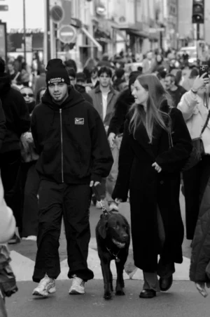 Thomas · 01/11/2025 · 1080 x 1625 Cette photographie en noir et blanc met en scène un couple marchant en ville. Le sujet principal est un homme en sweat-shirt et une femme en manteau, tenant un chien en laisse.La composition montre une rue animée avec une foule en arrière-plan. Le cadrage est serré sur le couple, mais inclut aussi des éléments urbains. La lumière semble naturelle, probablement en journée, avec des contrastes marqués. Les couleurs dominantes, bien que monochromes, évoquent des tons gris et noirs.Le style est documentaire, capturant un moment réel. L’ambiance est dynamique et urbaine, reflétant la vie quotidienne dans une ville.