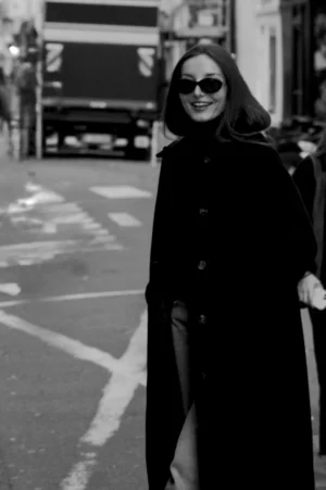 Thomas · 01/11/2025 · 1080 x 1625 Cette photographie en noir et blanc met en scène une femme comme sujet principal, vêtue d’un long manteau sombre et d’un chapeau, marchant sur un trottoir urbain. La composition est simple et équilibrée, avec un cadrage serré sur elle, légèrement décentré.La lumière, naturelle et douce, crée des contrastes subtils sur son visage et ses vêtements. L’ambiance est à la fois élégante et intemporelle, renforcée par le style rétro des années 1950-60. Les couleurs dominantes, bien que monochromes, jouent sur les nuances de gris et de noir, accentuant le côté sophistiqué et mystérieux de la scène.