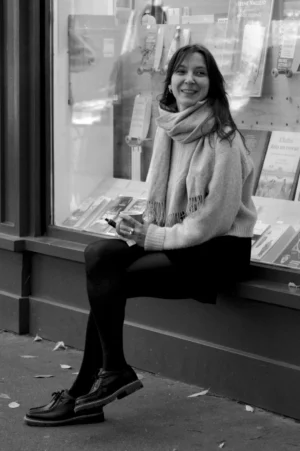 Thomas · 01/11/2025 · 1080 x 1625 Cette photographie en noir et blanc met en scène une jeune femme comme sujet principal, assise sur un rebord de fenêtre. Elle sourit en tenant un livre ou un magazine.La composition est simple et naturelle, avec un cadrage serré sur elle, mais laissant voir une partie de la rue et des affiches derrière elle. La lumière est douce et semble naturelle, éclairant son visage et créant des contrastes subtils.L’ambiance est chaleureuse et détendue, évoquant un moment de pause ou de lecture paisible en ville. Le style est documentaire et spontané.