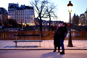 Cette photographie capture un couple en train de s'embrasser près d'un pont, sous un réverbère allumé. Le sujet principal est ce moment intime, mis en valeur par un cadrage serré.La composition utilise une perspective légèrement basse, avec le pont et les bâtiments en arrière-plan. La lumière douce et chaude des réverbères crée une ambiance romantique et tamisée. Les couleurs dominantes sont les tons chauds (orange, beige) contrastant avec les teintes froides des bâtiments et de la nuit.Le style est réaliste et poétique, évoquant une scène urbaine nocturne paisible et émouvante.