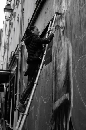 Philippe · 01/11/2025 · 2001 x 3000 Cette photographie en noir et blanc montre un homme en train de peindre ou d’appliquer une substance sur une façade verticale à l’aide d’un rouleau, depuis un échafaudage.Le **sujet principal** est cet homme, dont le travail est mis en valeur par un **cadrage serré**. La **composition** est dynamique, avec une perspective ascendante renforcée par les barres de l’échafaudage. La **lumière**, contrastée, accentue les ombres et les textures, créant une ambiance industrielle et urbaine. Le **style** est documentaire et réaliste, évoquant un travail manuel. L’absence de couleurs renforce l’aspect brut et intemporel de la scène.