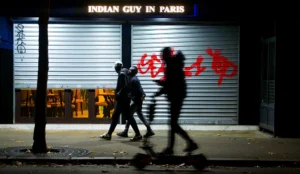 Cette photographie capture trois silhouettes floues en mouvement devant un magasin nommé **« Indian Guy in Paris »**. Le cadrage est serré, mettant l’accent sur les figures et les enseignes lumineuses.La lumière est tamisée, avec des teintes chaudes (jaunes et oranges) provenant des vitrines éclairées. Les couleurs dominantes sont les tons sombres de la nuit, contrastant avec les éclats colorés des enseignes.Le style est réaliste et dynamique, évoquant une ambiance nocturne urbaine, presque mystérieuse et vivante.