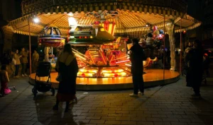 Cette photographie capture un **manège illuminé** la nuit, sujet principal. La composition montre des enfants et des adultes autour, avec un cadrage serré sur la structure colorée et ses lumières vives.Les jeux de lumière, chauds et artificiels, créent une ambiance festive et magique. Les couleurs dominantes sont les **tons chauds** (oranges, jaunes) et les éclats lumineux multicolores.Le style est **documentaire et nocturne**, évoquant une scène de fête foraine. L’ambiance est joyeuse et animée, malgré l’obscurité environnante.