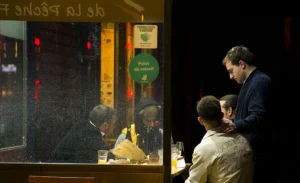 Cette photographie capture une scène nocturne dans un bar ou un restaurant. Le sujet principal est un groupe de personnes assises autour d'une table, avec un homme en costume penché vers elles.La composition montre un cadrage serré sur les individus, avec une lumière tamisée et des reflets sur la vitre. Les couleurs dominantes sont les tons chauds (jaunes, oranges) et les teintes sombres, créant une ambiance intimiste et conviviale. Le style est réaliste et documentaire, mettant en valeur une ambiance nocturne et urbaine.