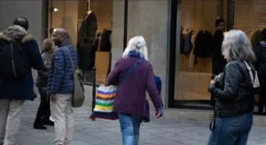 Muriel · 1080 x 592 Cette photographie montre un groupe de personnes en mouvement devant une entrée vitrée. Le sujet principal semble être une femme au centre, vêtue d’un pull violet et portant un sac coloré, marchant vers la gauche.La composition est dynamique, avec un cadrage serré sur les passants. La lumière est naturelle, tamisée, créant des ombres douces. Les couleurs dominantes sont sobres : tons neutres (gris, bleu) et quelques éclats vifs (sac multicolore).Le style est documentaire et réaliste, capturant une scène urbaine du quotidien. L’ambiance est calme et ordinaire, reflétant une journée banale en ville.