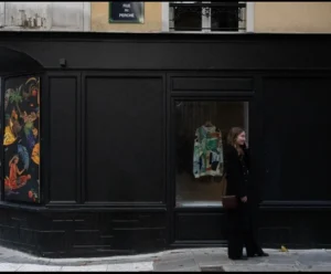 Muriel · 948 x 784 Cette photographie montre une personne en manteau noir entrant ou sortant d’une petite porte vitrée, encastrée dans un mur sombre.La composition est épurée, avec un cadrage serré sur la porte et le sujet. La lumière est tamisée, créant des contrastes doux et des ombres marquées. Les couleurs dominantes sont les tons sombres (noir, gris) avec des touches de jaune et de rouge sur les affiches murales.Le style est sobre et urbain, évoquant une ambiance mystérieuse et intimiste, presque cinématographique. L’atmosphère est calme et légèrement mélancolique.