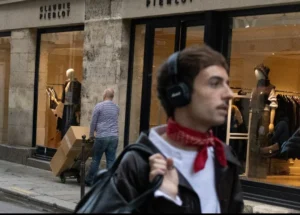 Muriel · 1080 x 773 Cette photographie met en scène un jeune homme comme sujet principal, marchant avec des écouteurs, un sac et un bandana rouge. La composition montre une rue urbaine avec des vitrines et des passants en arrière-plan.Le cadrage est serré sur le jeune homme, mais inclut des éléments contextuels. La lumière est naturelle, légèrement tamisée, créant des ombres douces. Les couleurs dominantes sont neutres (gris, beige) avec des touches de rouge et noir.Le style est réaliste et documentaire, capturant un instant de vie quotidienne. L’ambiance est urbaine et moderne, avec une touche de dynamisme et de modernité.