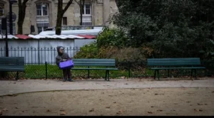 Muriel · 1080 x 597 Cette photographie met en scène une personne, sujet principal, de dos, vêtue d’un manteau bleu, tenant un sac. La composition montre un parc urbain avec des bancs verts alignés et une haie de buissons.Le cadrage est serré sur la personne, avec un arrière-plan légèrement flou. La lumière est naturelle, tamisée, créant des ombres douces. Les couleurs dominantes sont les tons verts (végétation), gris (bâtiments, sol) et bleu (vêtement).Le style est réaliste et sobre, évoquant une ambiance calme et mélancolique, presque introspective.