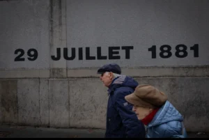 Muriel · 1080 x 723 Cette photographie montre deux personnes marchant devant un mur gris. Le **sujet principal** est la date "29 JUILLET 1881" gravée sur le mur, évoquant un lieu historique.La **composition** est simple, avec un **cadrage serré** sur les deux silhouettes en mouvement. La **lumière** est naturelle mais tamisée, créant des contrastes doux. Les **couleurs dominantes** sont les gris et bleus froids, renforçant une ambiance sobre et mélancolique.Le **style** est documentaire et sobre, avec une **ambiance** à la fois intemporelle et nostalgique, évoquant le passage du temps.