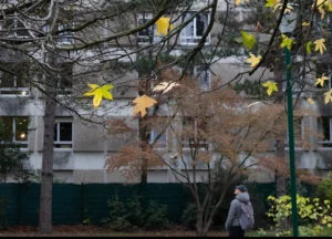 Muriel · 1080 x 777 Cette photographie met en scène une personne en mouvement, floue, marchant sous des arbres. Le sujet principal est suggéré par cette silhouette, presque effacée, créant un contraste avec l’environnement.La composition joue sur les plans superposés : les feuilles colorées en automne (jaunes, oranges) et les branches qui encadrent la scène. Le cadrage est serré, centré sur la personne et les arbres, avec un arrière-plan de bâtiments discrets.Les couleurs dominantes sont les tons automnaux chauds, contrastant avec le gris des murs. La lumière semble naturelle, tamisée, renforçant une ambiance paisible et mélancolique. Le style est réaliste et légèrement flou, évoquant une atmosphère contemplative et intime.