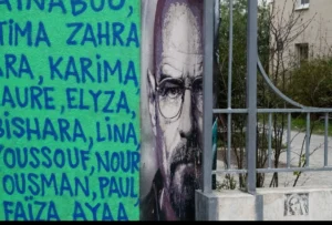 Muriel · 1080 x 730 Cette photographie montre un portrait mural d’un homme barbu, probablement Che Guevara, avec des noms féminins écrits en arabe à gauche. La composition inclut aussi une structure métallique et des plantes en arrière-plan.Le cadrage est serré sur le visage et les inscriptions. La lumière semble naturelle, avec des ombres douces. Les couleurs dominantes sont les tons verts, gris et noirs, créant une ambiance sobre et engagée. Le style est réaliste et graphique, évoquant un message politique et social. L’ambiance est à la fois sobre et puissante, soulignant un contexte militant ou commémoratif.