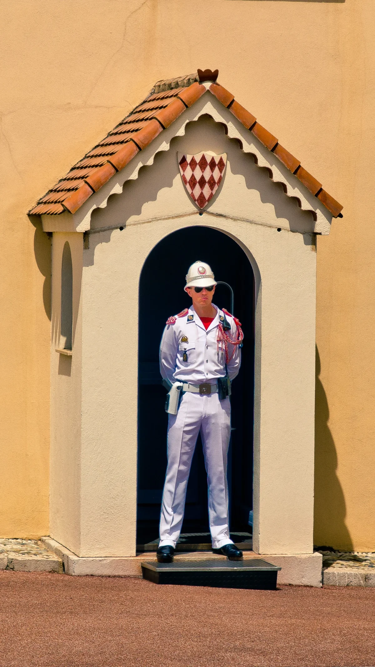 Tery · 1562 x 2777 Cette photographie montre un garde en uniforme blanc et casquette blanche, posté dans une niche architecturale ornée d’un toit en tuiles rouges et d’un blason. Le sujet principal est le garde, figé dans une posture rigide.La composition est symétrique, avec un cadrage serré mettant en valeur le personnage. La lumière naturelle éclaire frontalement la scène, créant des contrastes nets. Les couleurs dominantes sont le blanc de l’uniforme, le rouge des tuiles et du blason, et le beige de la niche.Le style est réaliste et photographique, évoquant une ambiance solennelle et formelle, typique d’un lieu historique ou officiel.