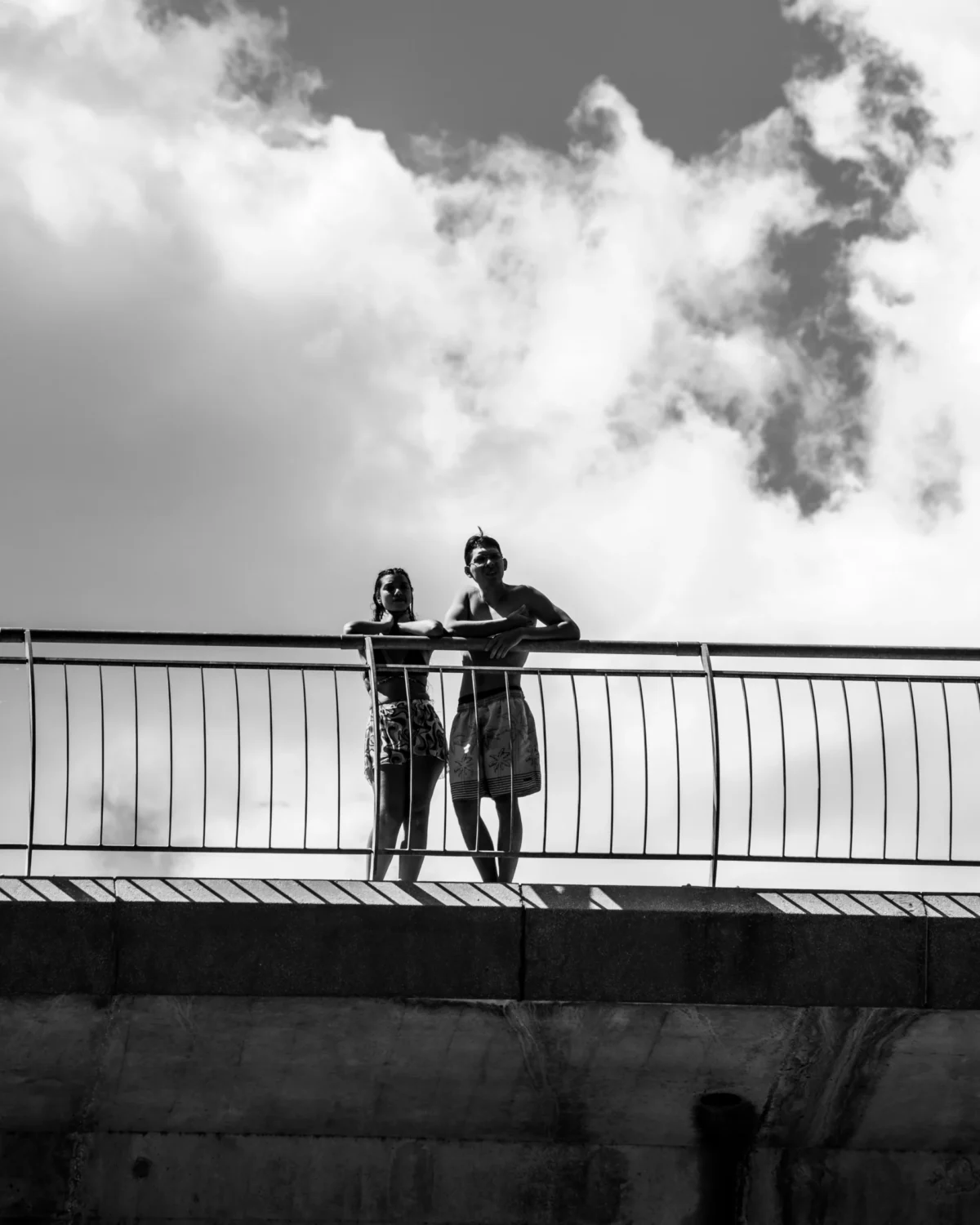 Tery · 4480 x 5600 Cette photographie met en scène deux personnes debout sur une passerelle métallique. Le sujet principal est ce couple, l’un derrière l’autre, en position intime et protectrice.La composition est épurée, avec un cadrage serré sur les sujets et un arrière-plan flou de ciel nuageux. La lumière, douce et naturelle, crée des contrastes marqués, accentuant les silhouettes.En noir et blanc, les tons dominants varient du clair au foncé, renforçant l’émotion. Le style est sobre et poétique, évoquant une ambiance à la fois mélancolique et tendre.