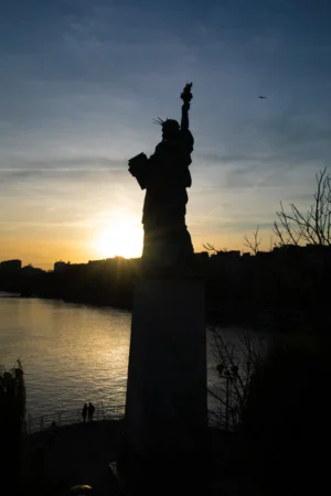 Cette photographie met en scène la **Statue de la Liberté**, silhouette principale, en contre-jour. La composition est épurée, avec un cadrage serré sur la statue, encadrée par des branches nues. La lumière dorée du coucher de soleil crée un effet dramatique, accentuant les contours et projetant une aura lumineuse. Les couleurs dominantes sont les **tons chauds (oranges, jaunes)** contrastant avec le bleu nuit du ciel et les ombres sombres. Le style est **sobre et poétique**, évoquant une ambiance **mélancolique et majestueuse**, renforcée par la sérénité du fleuve en arrière-plan.