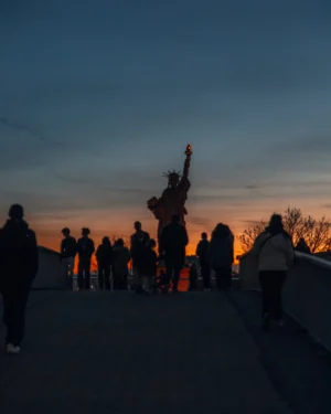 Cette photographie capture un groupe de personnes en silhouette lors d’un coucher de soleil. Le sujet principal semble être une personne levant les bras, entourée d’autres silhouettes en mouvement.La composition est dynamique, avec un cadrage serré sur les figures. La lumière douce et dorée du crépuscule crée des contrastes forts, accentuant les ombres. Les couleurs dominantes sont les teintes chaudes (orange, rouge) du ciel, contrastant avec les silhouettes sombres.Le style est épuré et naturel, évoquant une ambiance contemplative et presque mystique, renforcée par l’effet dramatique des ombres et de la lumière.