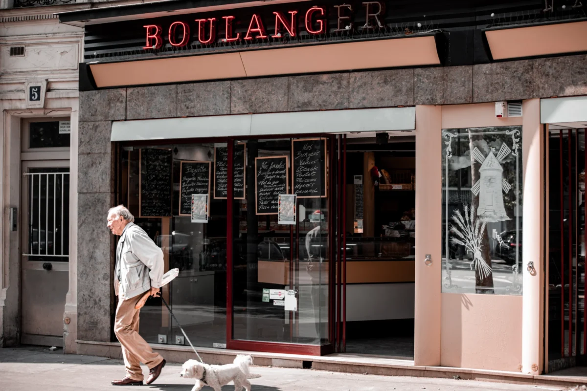Tery · 6627 x 4418 Cette photographie met en scène un boulanger en façade d'un commerce. Le sujet principal est un homme âgé marchant devant la boutique, portant un sac.La composition est équilibrée, avec un cadrage serré sur la devanture et le passant. La lumière est naturelle, douce et légèrement tamisée, créant des ombres douces. Les couleurs dominantes sont les tons neutres (beige, gris) avec des touches de rouge et noir.Le style est réaliste et documentaire, évoquant une ambiance calme et quotidienne, typique d’une rue urbaine paisible.