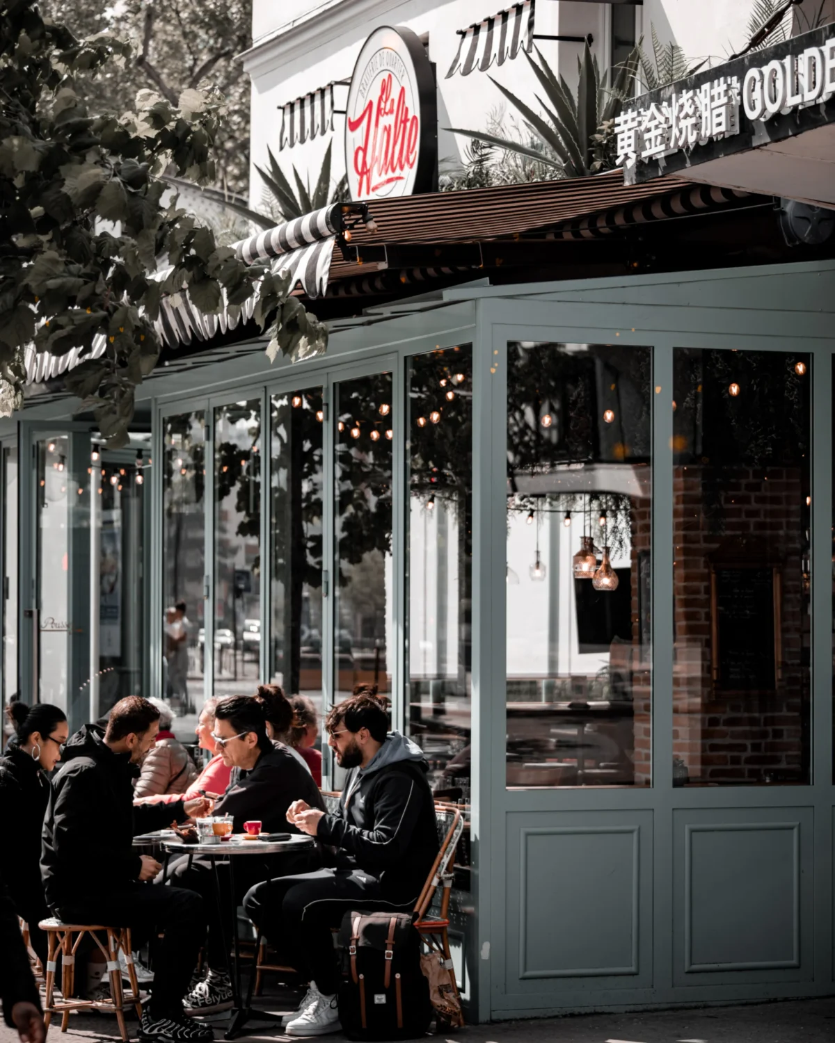 Tery · 4480 x 5600 Cette photographie montre un **restaurant extérieur** où le sujet principal est un groupe de personnes attablées, discutant et mangeant. La **composition** met en valeur une terrasse couverte avec des lumières chaudes et des décorations festives.Le **cadrage** est serré sur les convives, avec une profondeur de champ qui inclut aussi l’arrière-plan du restaurant. La **lumière** est douce et tamisée, créant une ambiance chaleureuse. Les **couleurs dominantes** sont des tons neutres (beige, gris) rehaussés par des éclairages dorés et des touches de rouge et vert.Le **style** est urbain et contemporain, évoquant une **ambiance conviviale et détendue**, presque intimiste malgré le cadre public.