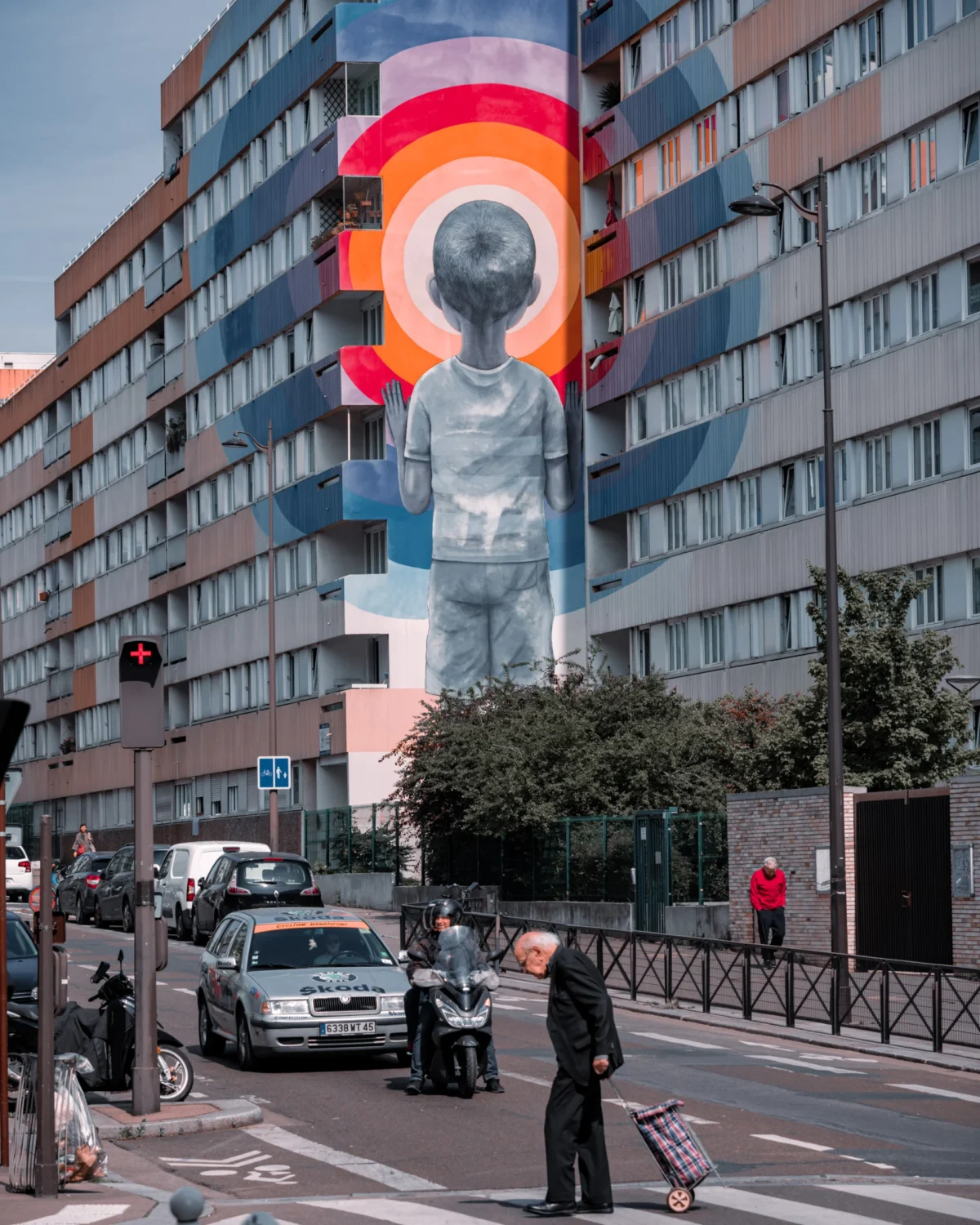 Tery · 4380 x 5475 Cette photographie met en scène un **mur géant couvert d'un street art** représentant un visage stylisé encadré par un arc-en-ciel. Le **sujet principal** est ce mural coloré, dominant l'arrière-plan urbain.La **composition** joue sur le contraste entre l'art moderne et l'architecture banale. Le **cadrage** inclut une rue animée avec des piétons et des voitures, ajoutant une touche de vie quotidienne.Les **couleurs dominantes** sont vives (rouge, bleu, jaune) pour le mural, tandis que les tons gris et beiges dominent le reste. La **lumière** est naturelle, claire, créant une ambiance **joyeuse et dynamique**, malgré un côté urbain réaliste.