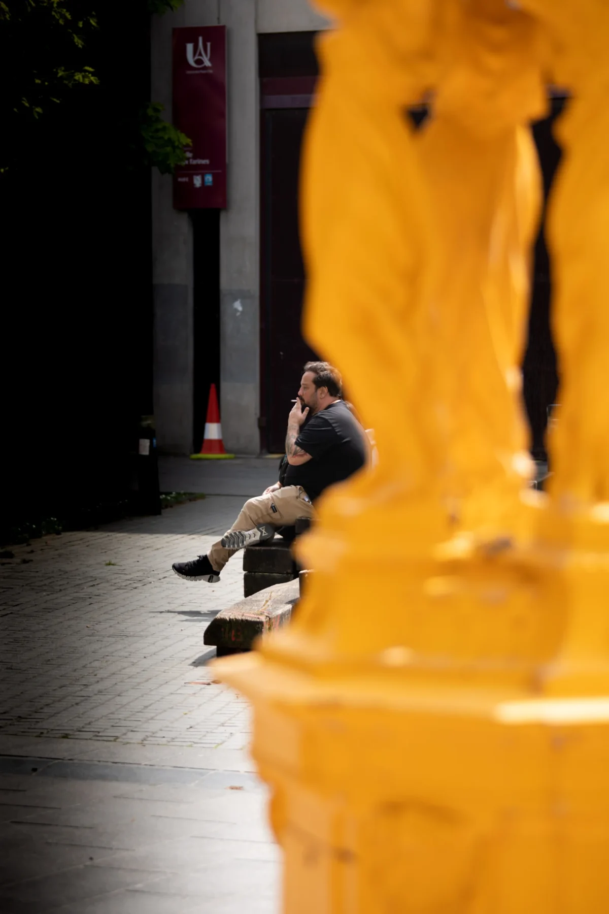 Tery · 4480 x 6720 Cette photographie met en scène un homme assis seul sur un banc, en silhouette partielle. La composition joue sur le contraste entre son ombre et la lumière vive. Le cadrage est serré, centré sur lui et un élément architectural jaune vif en premier plan, créant un effet de profondeur.Les couleurs dominantes sont les tons chauds (jaune, orange) et les ombres sombres. La lumière est naturelle et contrastée, avec des reflets et des contrastes marqués. Le style est réaliste et documentaire, évoquant une ambiance calme et mélancolique, presque urbaine et introspective.