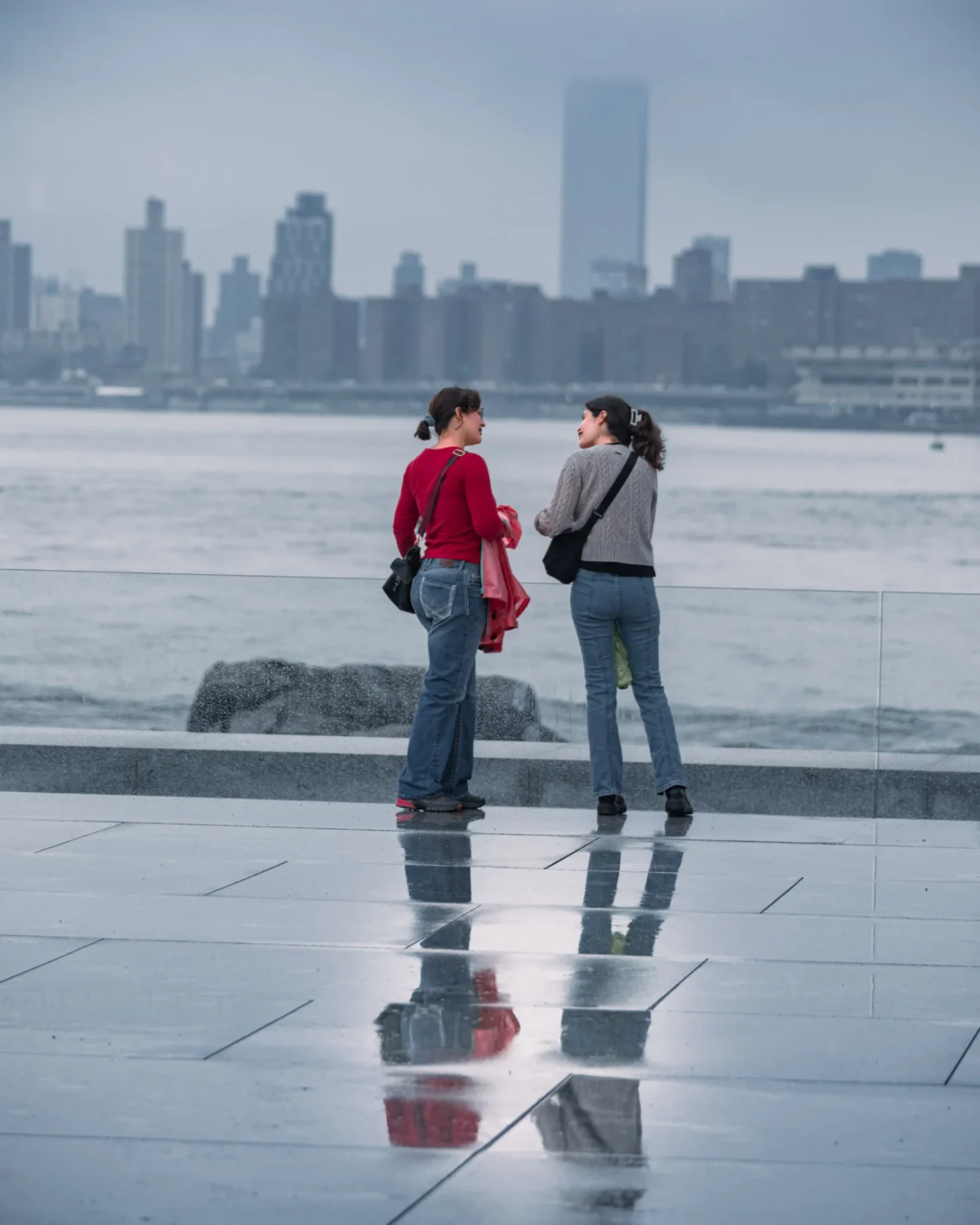 Tery · 4480 x 5600 Cette photographie met en scène deux personnes en conversation sur un rivage urbain. Le sujet principal est leur interaction, reflétée dans l’eau calme devant elles.La composition joue sur la symétrie et la réflexion, avec un cadrage serré sur les sujets et un arrière-plan de skyline floue. La lumière est douce, probablement en fin de journée, créant une ambiance paisible et légèrement mélancolique. Les couleurs dominantes sont les tons froids (bleu du ciel et de l’eau) et les teintes neutres, rehaussées par les vêtements rouges et bleus des personnages.Le style est épuré et contemplatif, évoquant une ambiance introspective et urbaine.