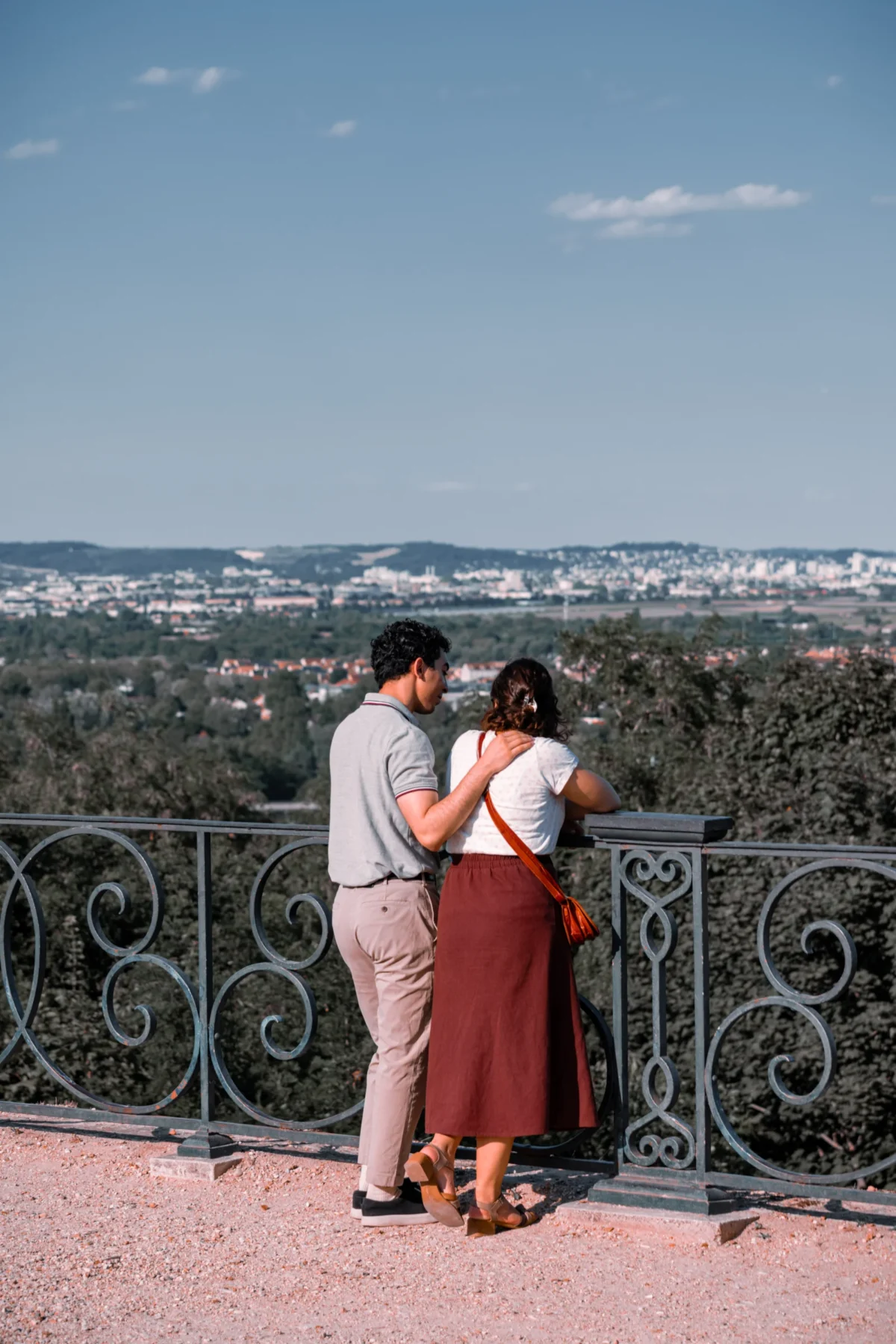 Tery · 4480 x 6720 Cette photographie met en scène un couple en embrassade sur une balustrade en métal ouvragé. Le sujet principal est leur complicité et leur connexion, renforcée par leur posture intimiste.La composition joue sur la profondeur avec une vue panoramique de la ville en arrière-plan. Le cadrage serré sur le couple, avec un arrière-plan flou, crée un effet de proximité et d'intimité.La lumière est douce et naturelle, typique d'une journée ensoleillée. Les couleurs dominantes sont les tons chauds (rouge de la robe, gris du métal) contrastant avec les bleus du ciel et les verts des arbres.Le style est romantique et poétique, évoquant une ambiance tendre et nostalgique.