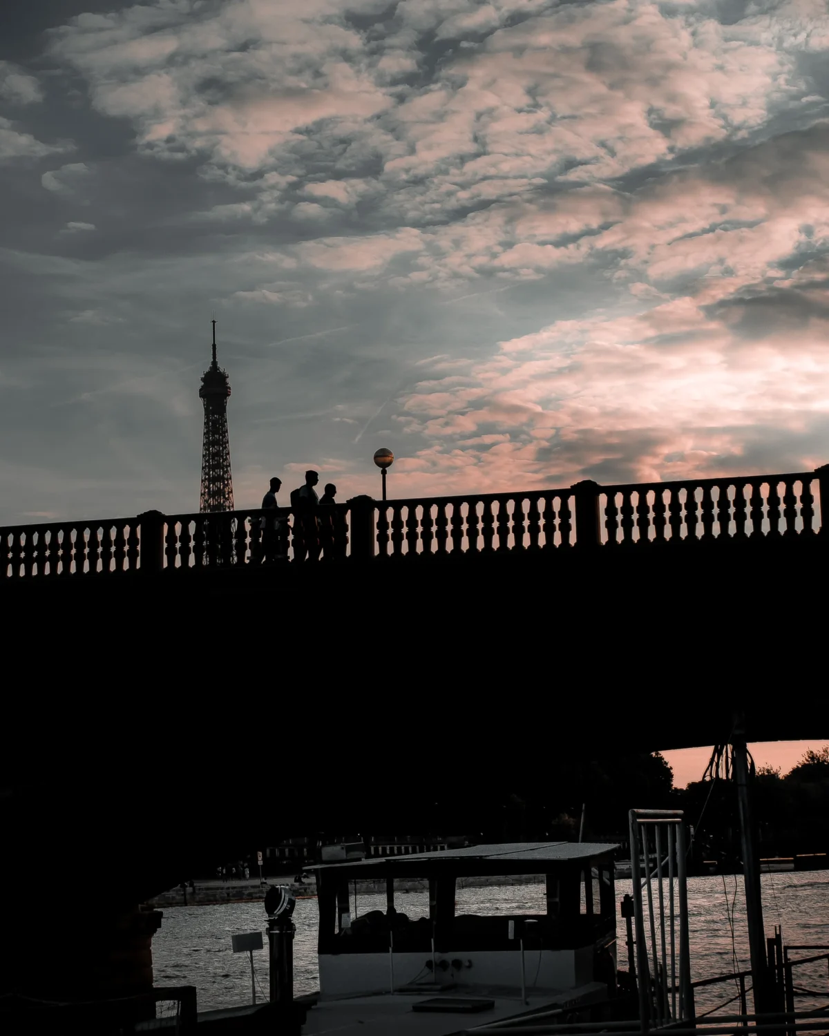Tery · 3648 x 4560 Cette photographie met en scène un **pont** avec une **barrière métallique** en premier plan. Le **sujet principal** est la silhouette d’une tour (probablement l’Eiffel) en arrière-plan, partiellement visible à travers les nuages.La **composition** joue sur les contrastes entre ombres et lumières, avec un **cadrage serré** sur le pont et une perspective légèrement basse. La **lumière** est douce, typique d’un coucher de soleil, créant une ambiance mélancolique. Les **couleurs dominantes** sont les gris, noirs et teintes rosées des nuages.Le **style** est épuré, presque monochrome, renforçant une **ambiance contemplative et nostalgique**.